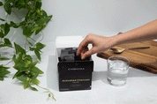 A hand reaches into a box labeled “Cymbiotika Activated Charcoal Daily Detox 26 Servings” by JING Organics, beside a glass of water, a wooden cutting board, and a green leafy plant on a white surface.