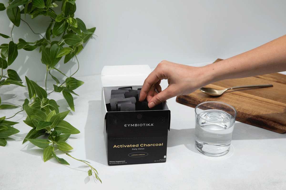 A hand reaches into a box labeled “Cymbiotika Activated Charcoal Daily Detox 26 Servings” by JING Organics, beside a glass of water, a wooden cutting board, and a green leafy plant on a white surface.