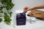 A hand reaches for a JING Organics Cymbiotika Liposomal Elderberry Defense 26 Servings packet from an open box beside a glass of water, gold spoon, wooden board, and green plant on a white surface.