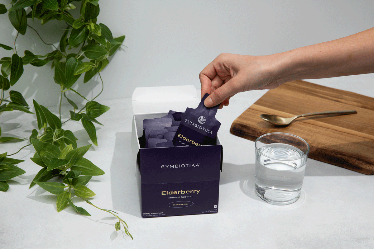 A hand reaches for a JING Organics Cymbiotika Liposomal Elderberry Defense 26 Servings packet from an open box beside a glass of water, gold spoon, wooden board, and green plant on a white surface.