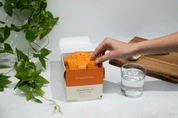 A hand reaches into an open box labeled "Cymbiotika Synergy Liposomal Vitamin C 30 Servings" by JING Organics, with orange packets visible. Nearby are a glass of water, a spoon on a wooden tray, and green leafy plants as decoration.