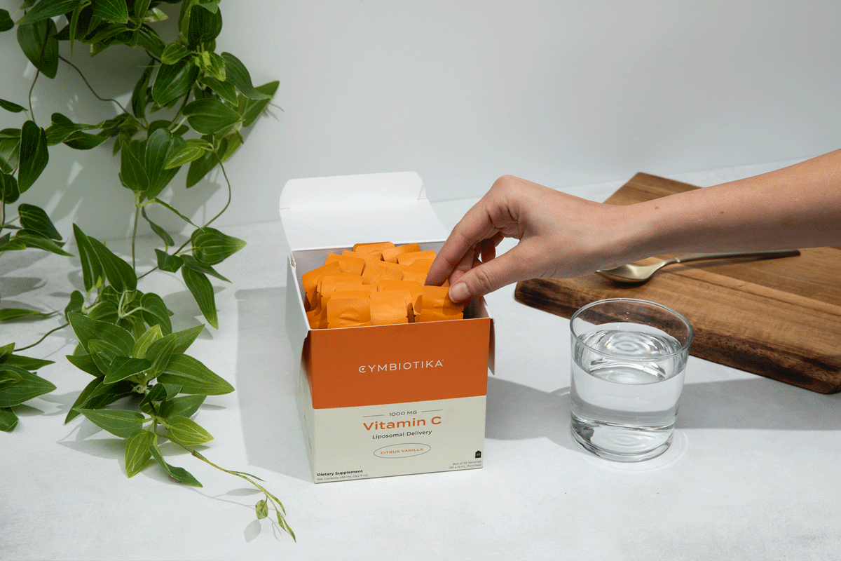 A hand reaches into an open box labeled "Cymbiotika Synergy Liposomal Vitamin C 30 Servings" by JING Organics, with orange packets visible. Nearby are a glass of water, a spoon on a wooden tray, and green leafy plants as decoration.