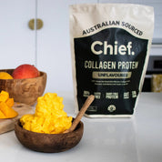 A wooden bowl with a yellow creamy mixture and spoon sits on a white countertop. Behind it are a bowl of mangoes and a bag labeled JING Organics Chief Collagen Protein Unflavoured 450g.