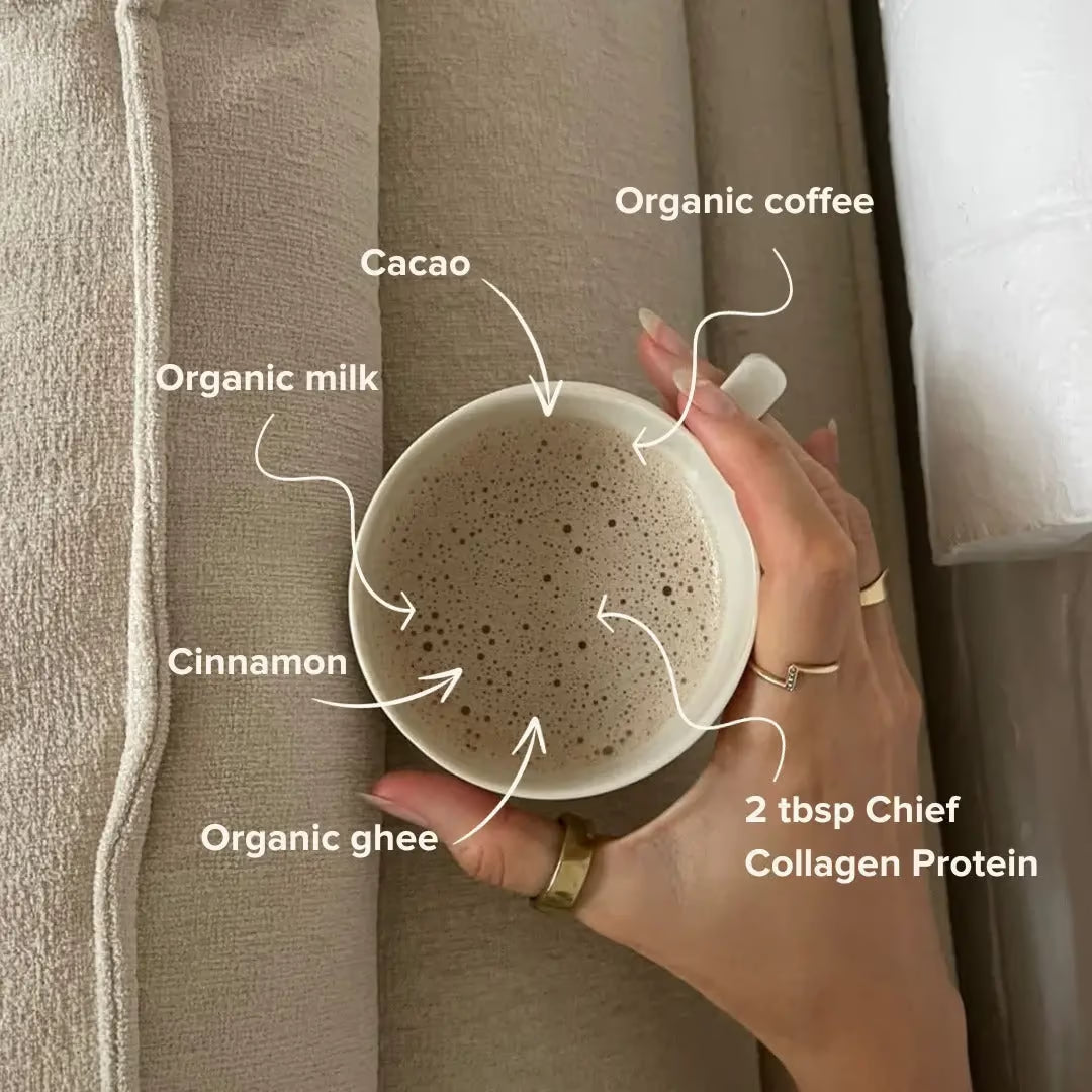 A hand holds a mug of frothy coffee with labeled ingredients: organic coffee, cacao, organic milk, cinnamon, organic ghee, and 2 tbsp JING Organics Chief Collagen Protein Creamy Vanilla 450g. The mug rests on a beige fabric surface.