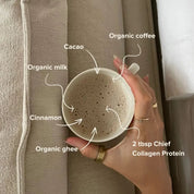 A hand holds a mug of frothy coffee with labeled ingredients: organic coffee, cacao, organic milk, cinnamon, organic ghee, and 2 tbsp JING Organics Chief Collagen Protein Creamy Vanilla 450g. The mug rests on a beige fabric surface.