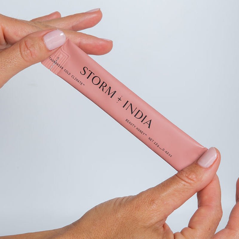 A person holds a pink sachet labeled JING Organics Storm + India Beauty Honey with both hands against a plain light background. The sachet is slender and rectangular.