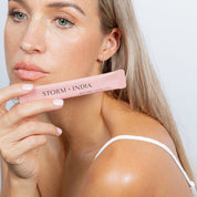 A woman with long blonde hair and natural makeup holds a pink tube of Storm + India Beauty Honey from JING Organics near her face. She wears a white spaghetti strap top and gazes thoughtfully to the side against a light background.