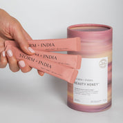 A hand holds three pink packets of Storm + India Beauty Honey by JING Organics beside a matching cylindrical container, all set against a plain white background.