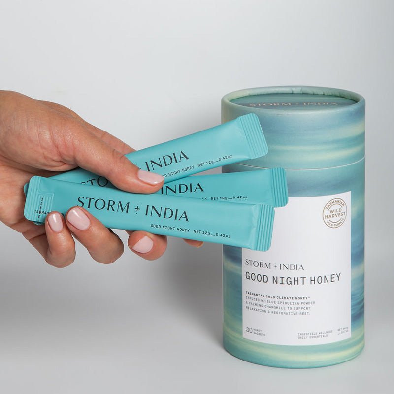 A hand holds two blue sachets labeled Storm + India Good Night Honey by JING Organics in front of a matching cylindrical container with the same branding and Wild Harvest label, set against a plain background.