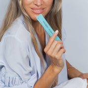 A woman with long, light brown hair and glossy lips holds a turquoise sachet of Storm + India Good Night Honey by JING Organics close to her face, wearing a light blue robe against a plain, light background.