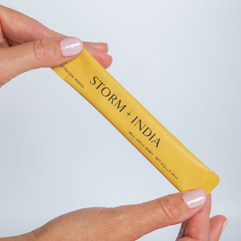 A person with light pink nail polish holds a yellow packet labeled JING Organics Storm + India Well Child Honey against a plain light background.