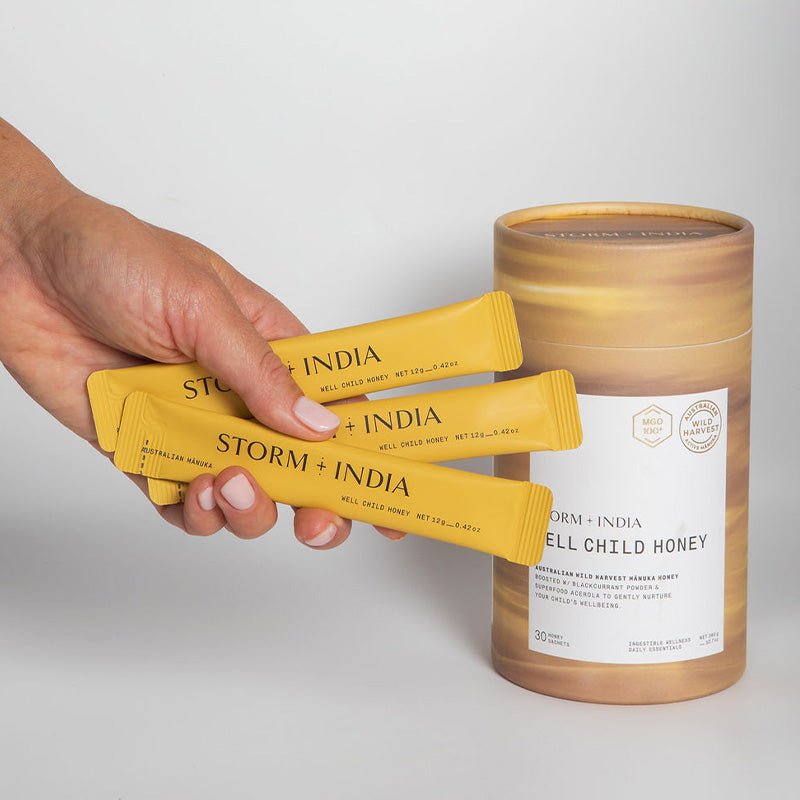 A hand holds three yellow JING Organics Storm + India Well Child Honey sachets beside a matching cylindrical container labeled with the product name, set against a plain white background.